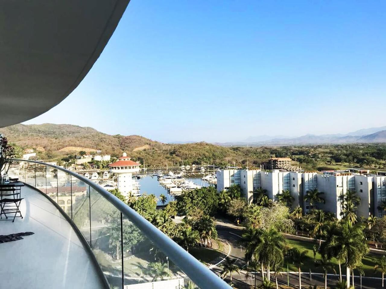 Ein Balkon mit gebogener Geländer, der auf einen Yachthafen, Palmen und Gebäude im Hintergrund blickt.