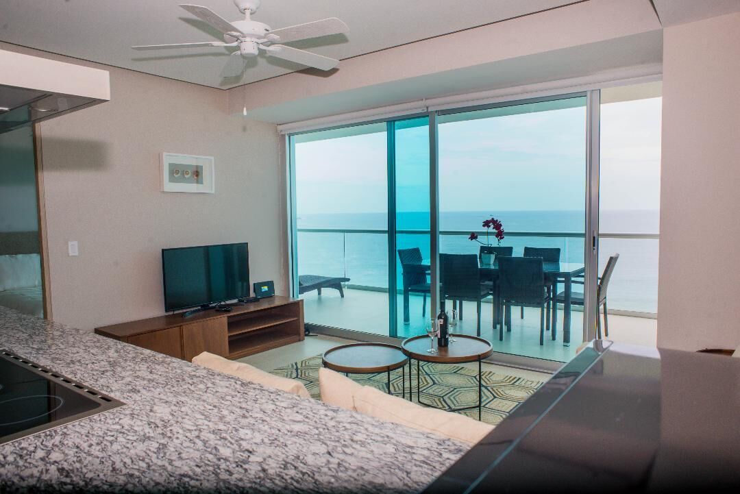 A modern living room with a TV, seating, and a balcony with a view of the ocean.