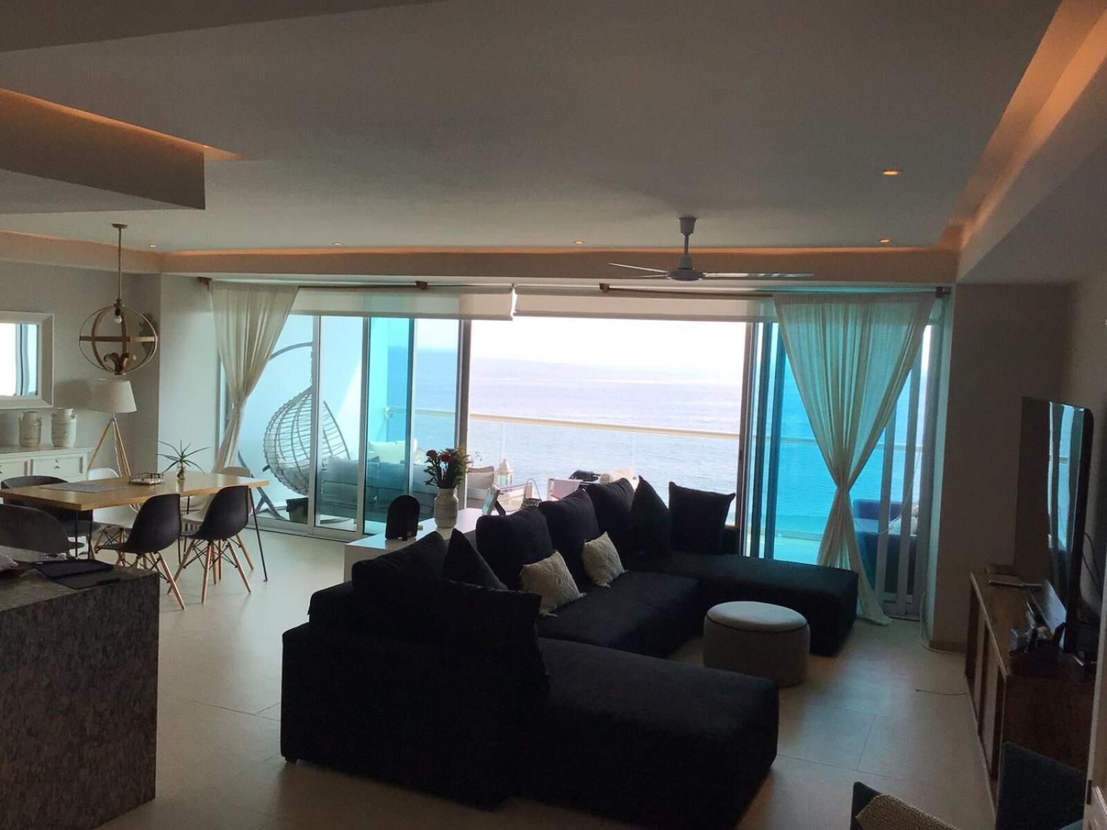 Uma sala de estar moderna com um sofá seccional preto grande, uma área de jantar com mesa e cadeiras, e vista para o oceano através de grandes janelas.