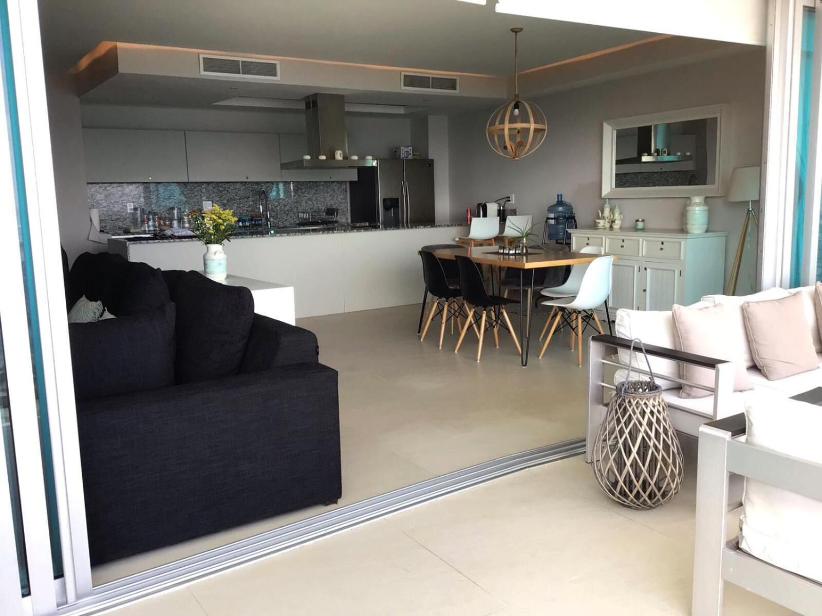 A modern living room with a black sofa, a dining area with a wooden table and chairs, and a kitchen in the background.