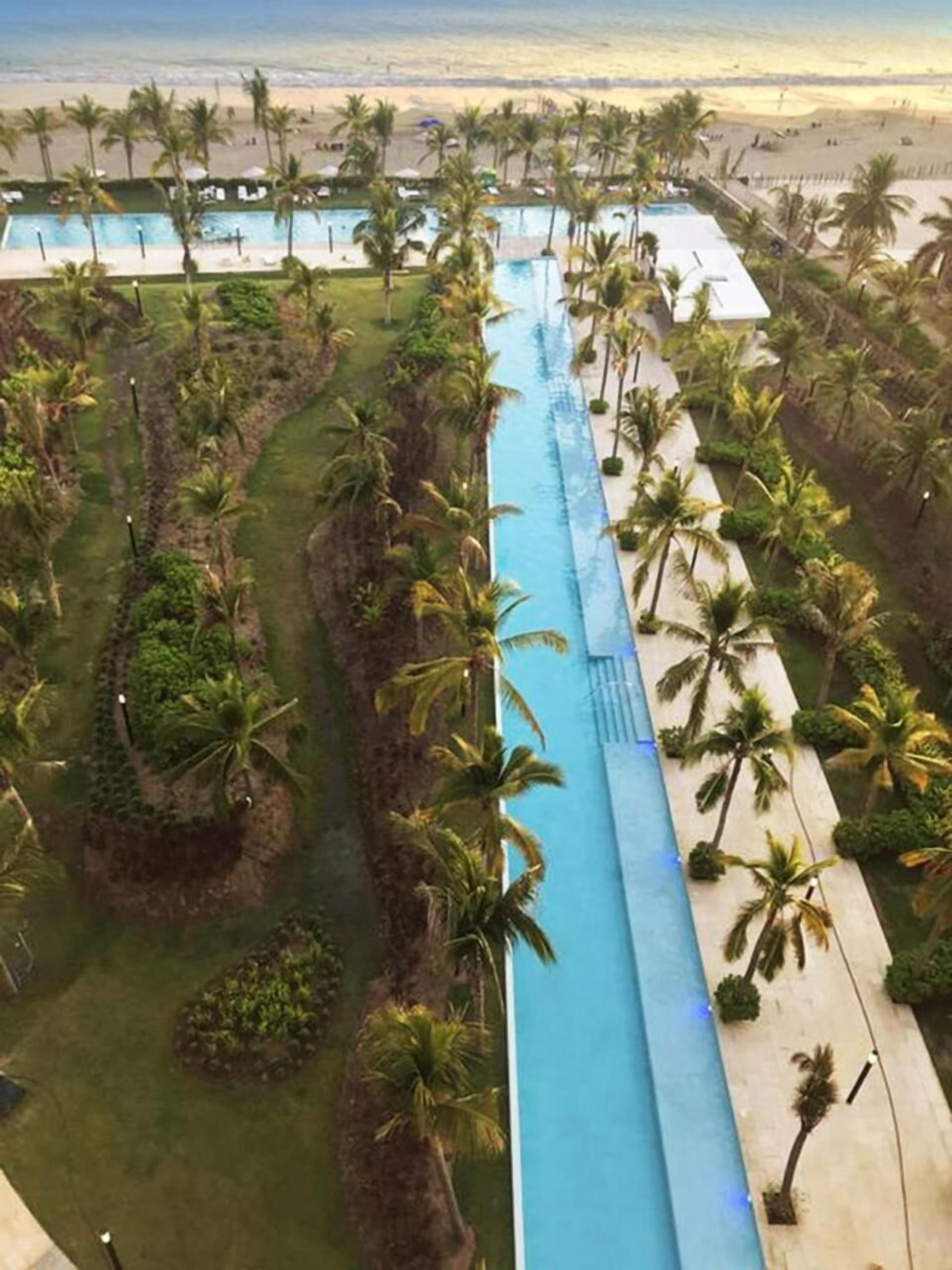 A long swimming pool with palm trees and a beach in the background.