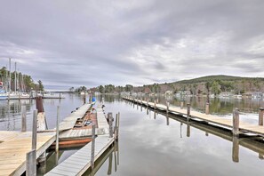 A calm waterfront with wooden docks extending into the water, surrounded by trees and houses. - Walk to Marina: Cute Gilford Cottage w/ Grill (Gilford)