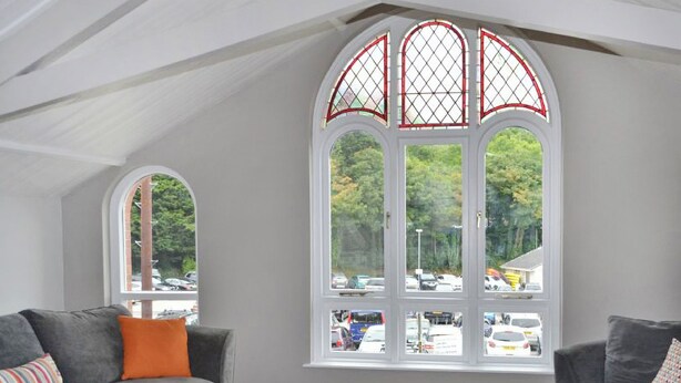 Ein modernes Wohnzimmer mit einem grauen Sofa, einem Holztisch und einem großen Bogenfenster mit Blick auf einen Parkplatz und Bäume.