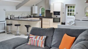 Eine moderne Küche mit weißen Schränken, einer zentralen Kochinsel und einem Essbereich mit grauem Sofa und bunten Kissen.