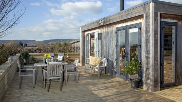 En træterrasse med udendørs møbler, udsigt over landskabet og en moderne bygning med glasdøre.