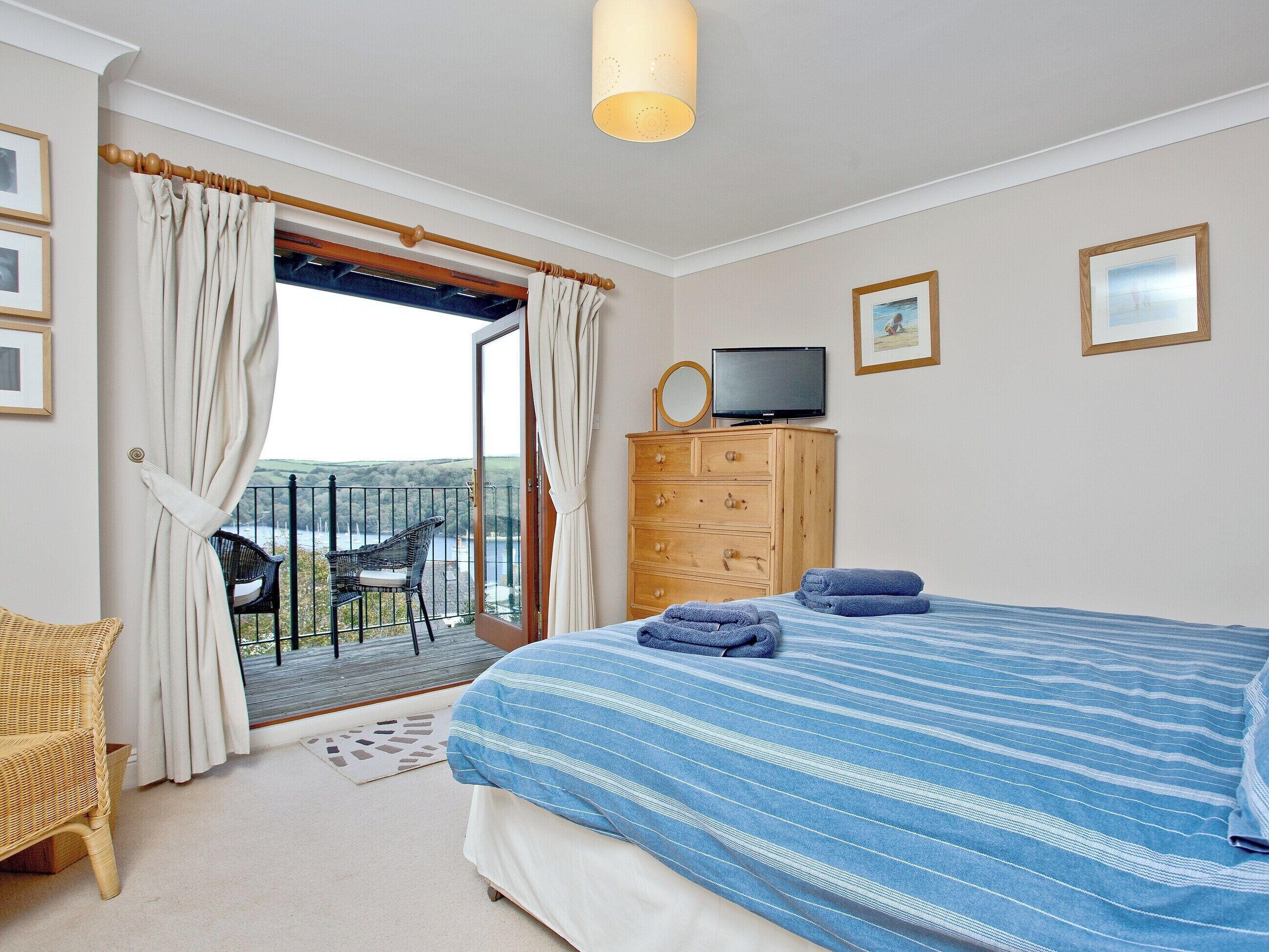 A bedroom with a bed, a wooden dresser, a television, and a balcony with a chair.