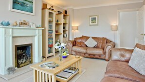 A cozy living room with a fireplace, leather sofa, wooden coffee table, and a bookshelf filled with books and decorative items. - 3 bedroom accommodation in Fowey (Fowey)