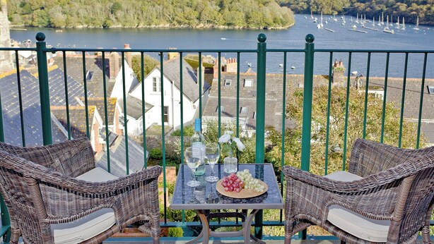 A balcony with wicker furniture, a table with wine glasses and grapes, and a view of a river with boats and houses in the background. - 3 bedroom accommodation in Fowey (Fowey)
