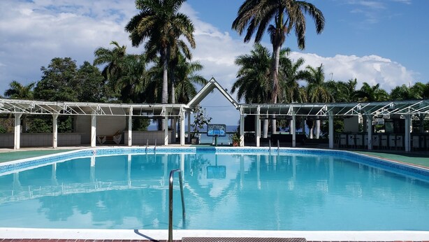 A swimming pool with a clear blue sky, palm trees, and a covered seating area. - Hideaway  by the Sea (Montego Bay)