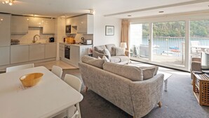 A modern kitchen with white cabinets, a dining table with a wooden bowl, a grey sofa, and a balcony with a view of the water. - 2 bedroom accommodation in Fowey (Fowey)