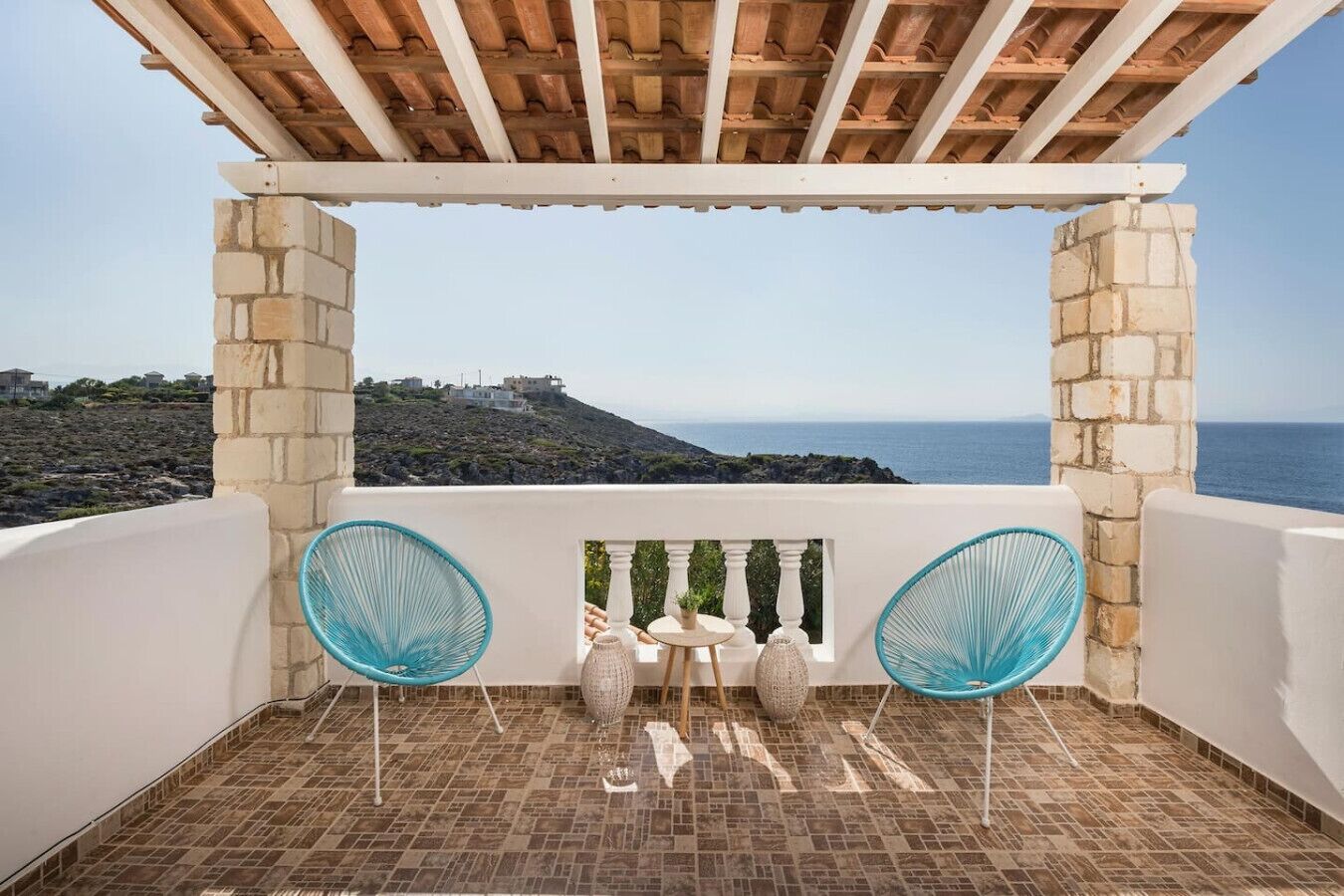 A balcony with a view of the sea, two blue chairs, a small table, and a stone pillar.
