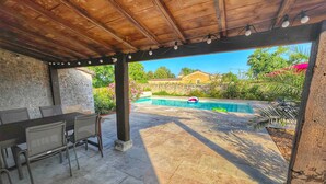 An outdoor patio with a pool, stone flooring, and a covered seating area with tables and chairs.