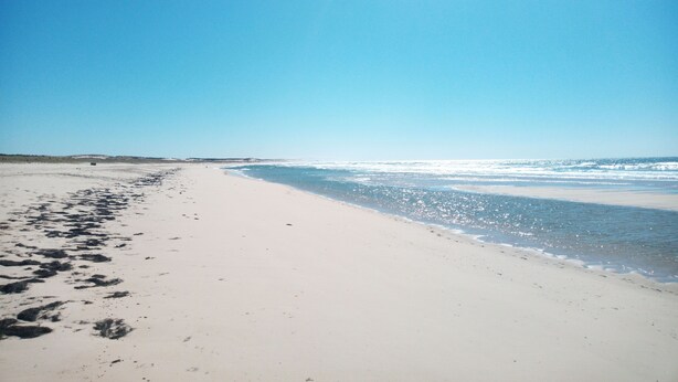 A wide, sandy beach with footprints leading to the water, under a clear blue sky.