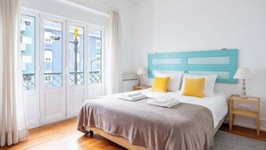 Et hotelværelse med en seng, et natbord, en lampe og udsigt til bygninger gennem et vindue.