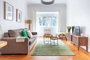 Olohuoneessa on sohva, kaksi sivupöytää, vihreä matto, televisio ja ikkuna, josta on näkymä rakennuksiin.