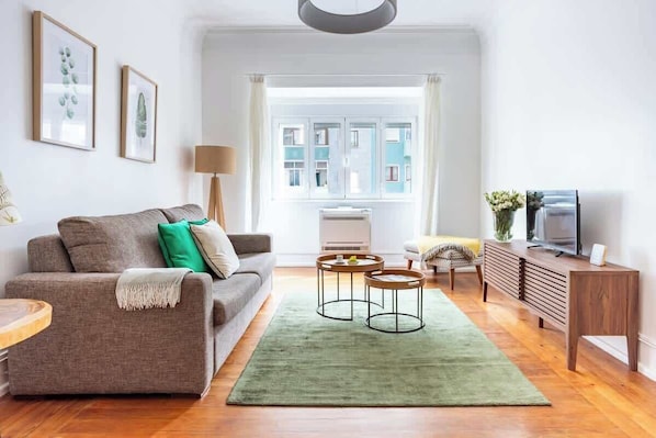 A living room with a sofa, two side tables, a green rug, a television, and a window with a view of buildings.
