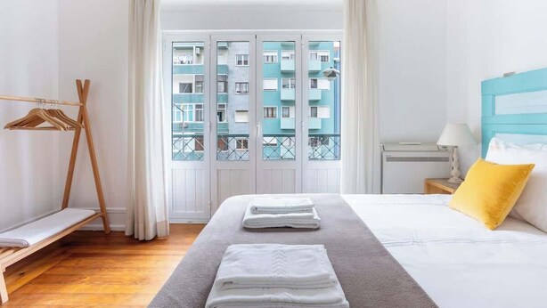 Et hotelværelse med en seng, et tøjholder af træ, et vindue med udsigt til en bygning og et natbord med en lampe.