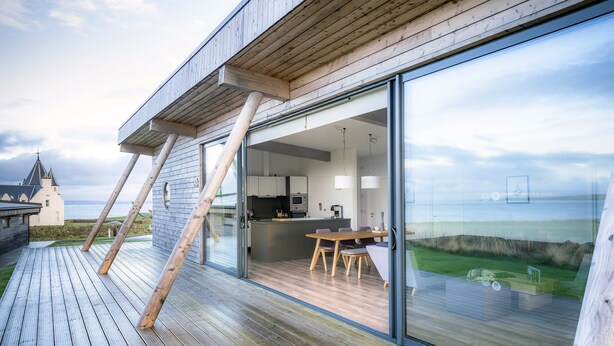 A modern house with a wooden deck, a dining area with a table and chairs, and a kitchen visible through glass doors.