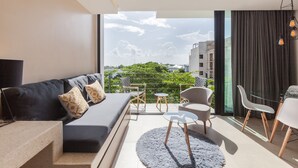 A modern living room with a sofa, chairs, a glass table, and a view of greenery and buildings through large windows. - Riva Playasur 405 - luxury studio - (playa del carmen)
