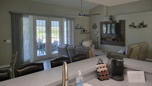 A modern living room with a sofa, coffee table, and a TV mounted on the wall. There is a dining area with a table and chairs, and a kitchen area with a coffee maker and a paper on the counter. - 3 bed/3 bath with Stunning Lakeview from private pool (Rotonda West)
