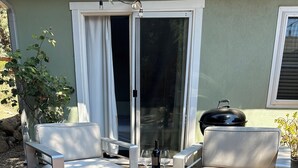 Un patio con dos sillas blancas acolchadas, una mesita, una botella de vino y una parrilla.
