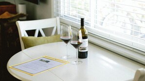 Una mesa blanca redonda con una botella de vino y una copa, frente a una ventana con persianas.