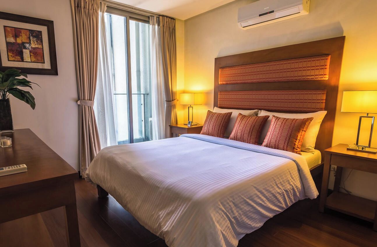 A hotel room with a bed, bedside lamps, a desk with a remote control, a potted plant, and an air conditioning unit.