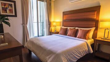 A hotel room with a bed, bedside lamps, a desk with a remote control, a potted plant, and an air conditioning unit.