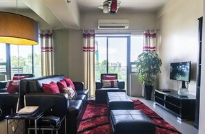 Een moderne woonkamer met een bank, voetenbankje en een tv op een meubel. Er is een groot raam met gordijnen, een potplant en een plafondlamp.
