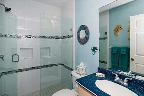 Ein modernes Badezimmer mit Duschkabine aus Glas, weißer Toilette, blauem Waschbecken und einer fleckigen Arbeitsplatte. Dekorative Wandbilder zeigen ein Seepferdchen und einen runden Spiegel.