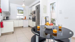 Een moderne keuken met witte kasten, een zwart glazen tafel en een fruitschaal met glaasjes sinaasappelsap.