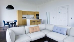 Uma sala de estar moderna com um sofá seccional, uma mesa de jantar com cadeiras e uma área de cozinha ao fundo.
