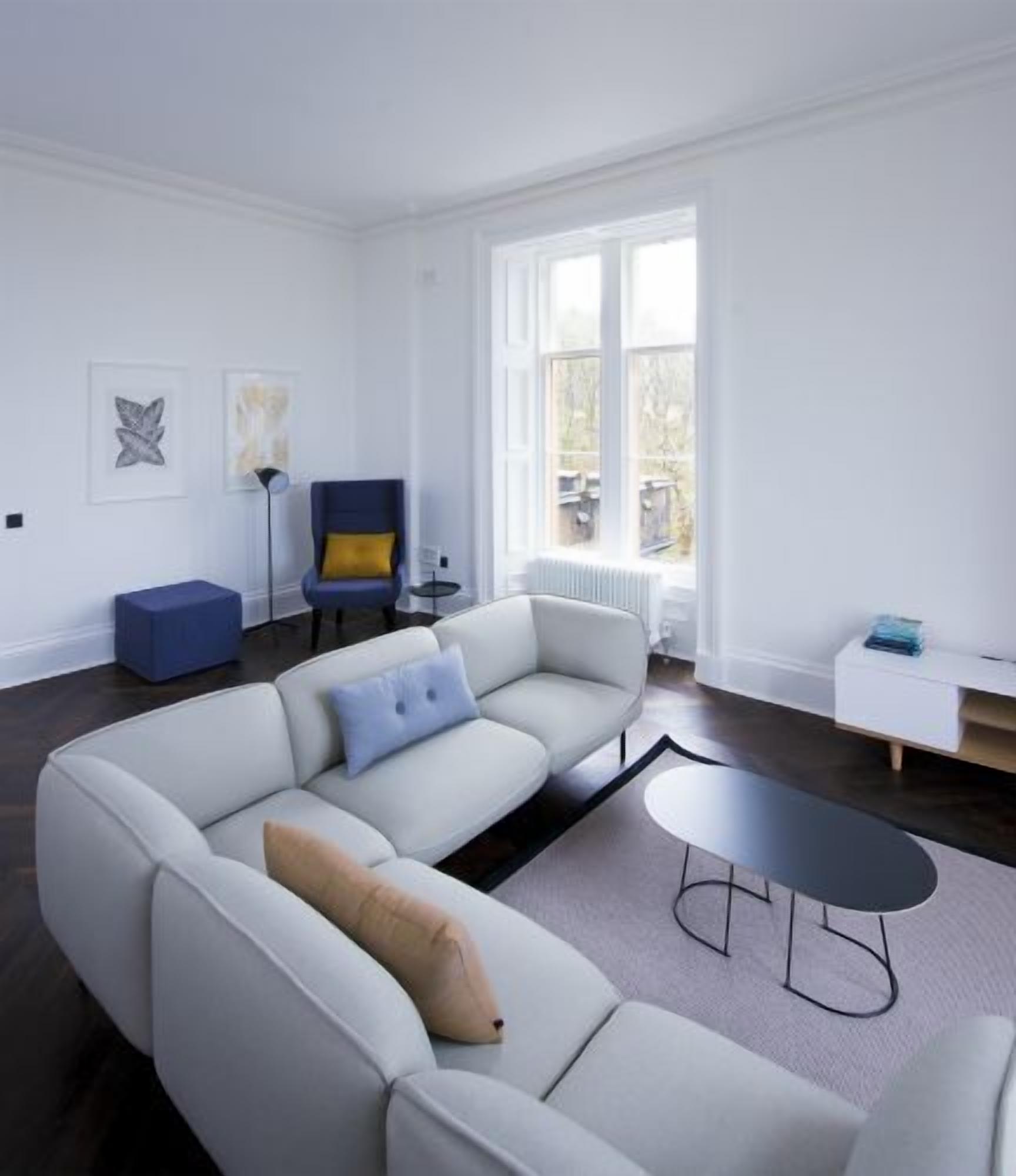 Un salón moderno con un amplio sofá en forma de L, una pequeña mesa redonda y un sillón azul. Hay una lámpara de pie y un radiador cerca de la ventana.