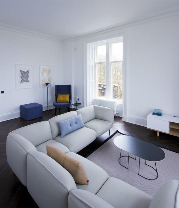 A modern living room with a large L-shaped sofa, a small round table, and a blue armchair. There is a floor lamp and a radiator near the window.