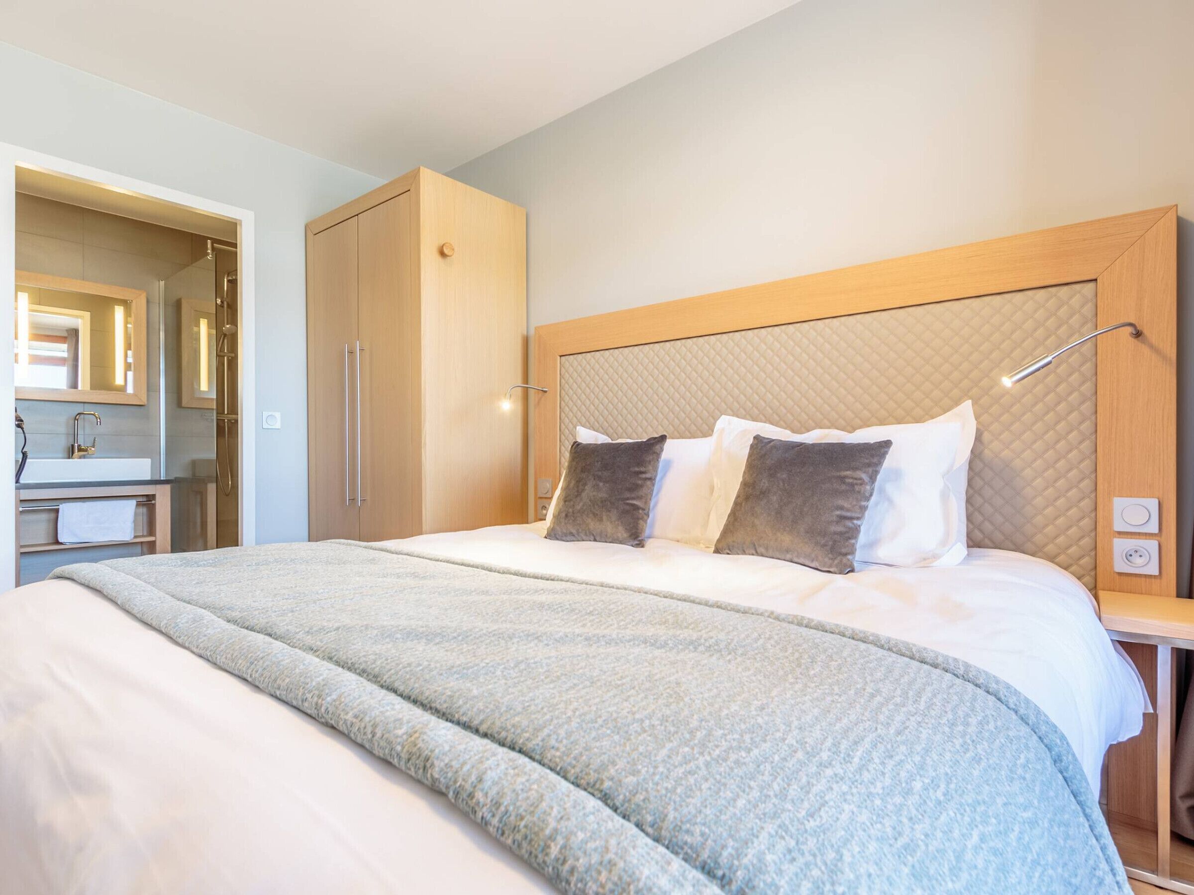 A hotel room with a large bed, a wooden wardrobe, a bedside table with a lamp, and a bathroom with a mirror and sink.