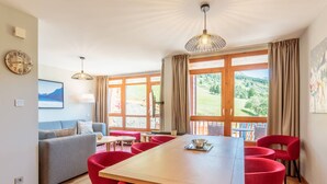 A modern dining area with a wooden table and red chairs, large windows with curtains, and a view of a green landscape.
