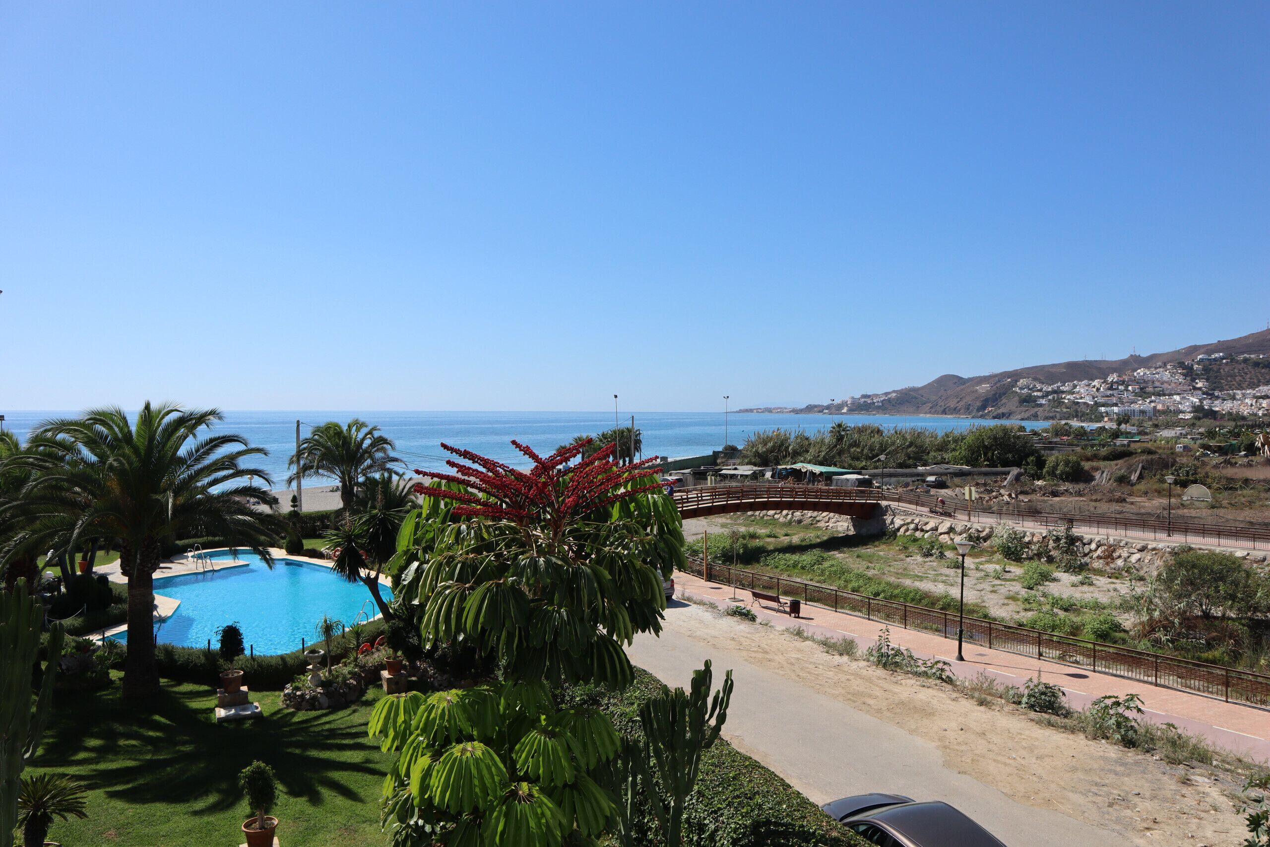 Una piscina rodeada de vegetación exuberante, un cielo azul despejado y una vista distante de un pueblo costero.