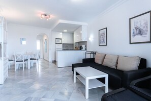 A modern living room with a black sofa and a white coffee table, a dining area with a white table and chairs, and a kitchen with white cabinets and appliances. - Stella Maris Beachfront 1B by Casasol (Nerja)