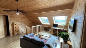 A cozy loft-style room with a wooden interior, a sloped ceiling fan, a desk with a computer, a sofa, and a large window offering a view.
