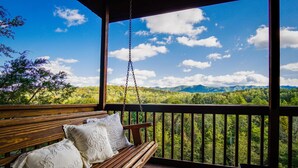 Una terraza de madera con un banco y cojines, con vistas a una cadena montañosa boscosa.