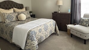 Una camera da letto con un letto dotato di testiera trapuntata, biancheria da letto fantasia, un comodino, una cassettiera, una sedia e una lampada fissata al muro.