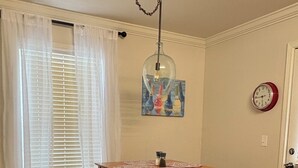Zona pranzo con tavolo e panche in legno, lampadario a sospensione, quadro, orologio e finestra con persiane.