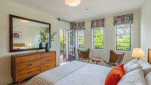 A bedroom with a large bed, wooden dresser, and a mirror. There are two windows with colorful curtains, a balcony with chairs, and a view of greenery. - Iconic, Unique Home in the Heart of Downtown SB! (Santa Barbara)