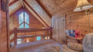 Um quarto de cabana acolhedor, com teto inclinado, uma janela grande, uma estante de livros e um espaço de estar confortável.