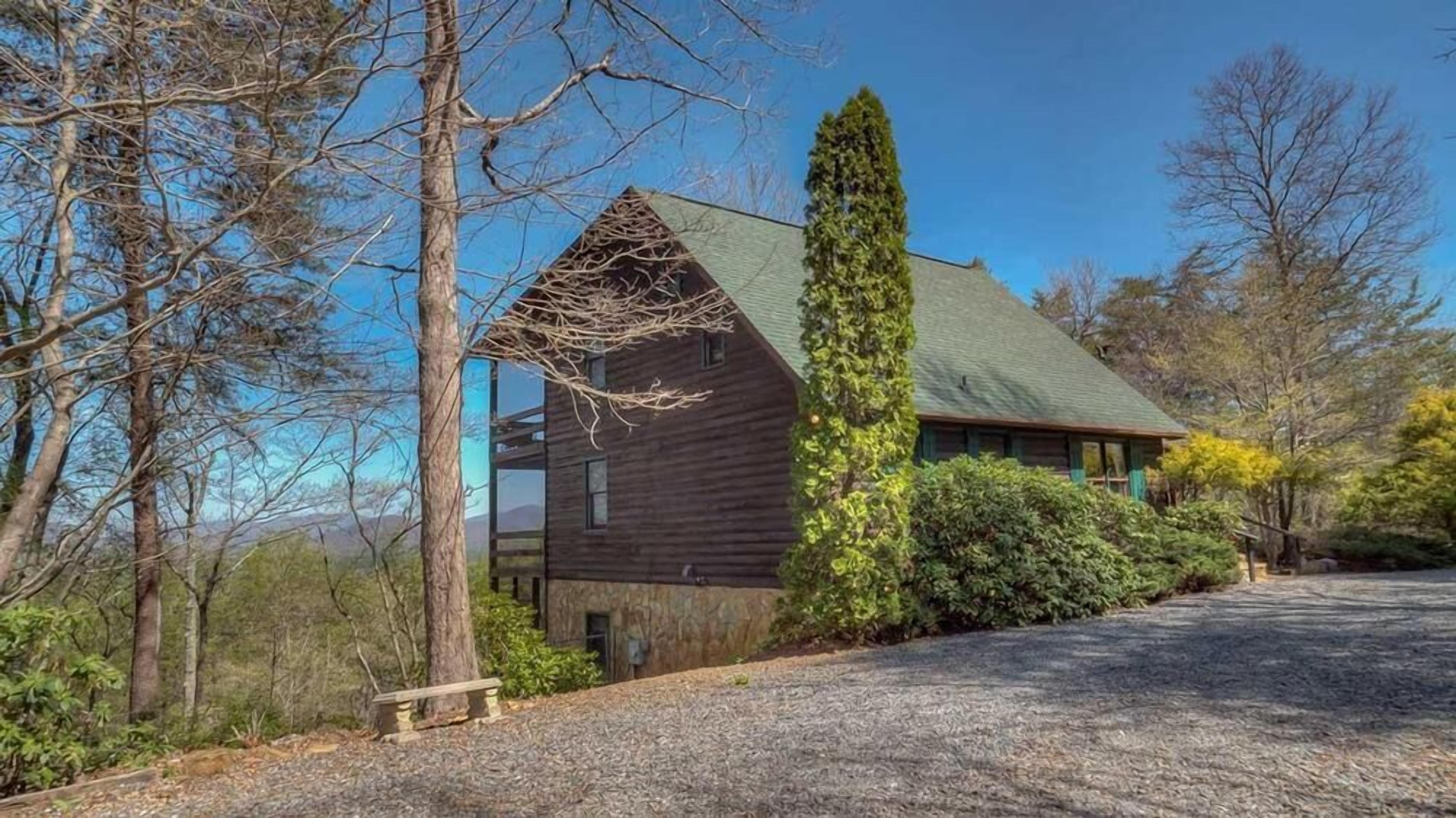 A wooden cabin with a sloped roof, surrounded by trees and a gravel driveway.