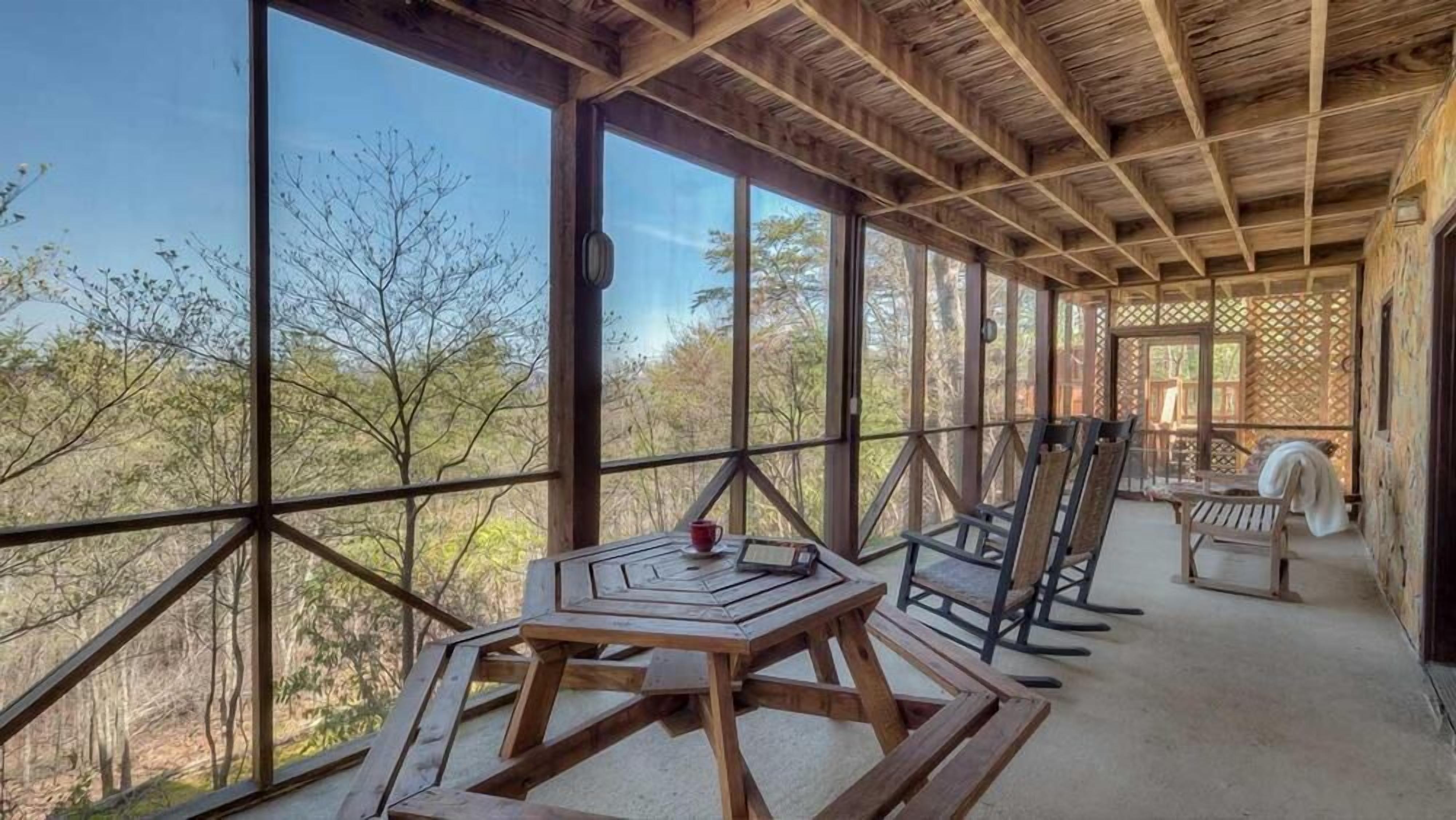 Un terrazzo in legno con tavolo e sedie, ampie finestre che si affacciano su alberi e una struttura a traliccio.
