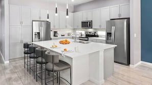 A modern kitchen with white cabinetry, stainless steel appliances, a central island with bar stools, and pendant lighting. - Fun Home Near Disney w/ Princess & Space BRs, Arcade, Game Loft, Theater & Pool (Davenport)