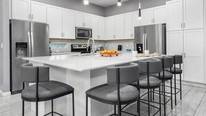 A modern kitchen with white cabinets, stainless steel appliances, a central island with bar stools, and pendant lighting. - Cozy Home Near Parks w Toy & BBall Theme BRs, Theater, Arcade, Game Loft, Pool (Davenport)