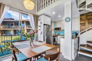 A modern kitchen with a stainless steel refrigerator, microwave, and oven. There is a dining area with a wooden table and chairs, and a balcony with a view of the outside.