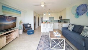 Un salon moderne avec un téléviseur, un canapé et une table basse. Il y a également un coin cuisine avec des meubles blancs et une table à manger avec des chaises.
